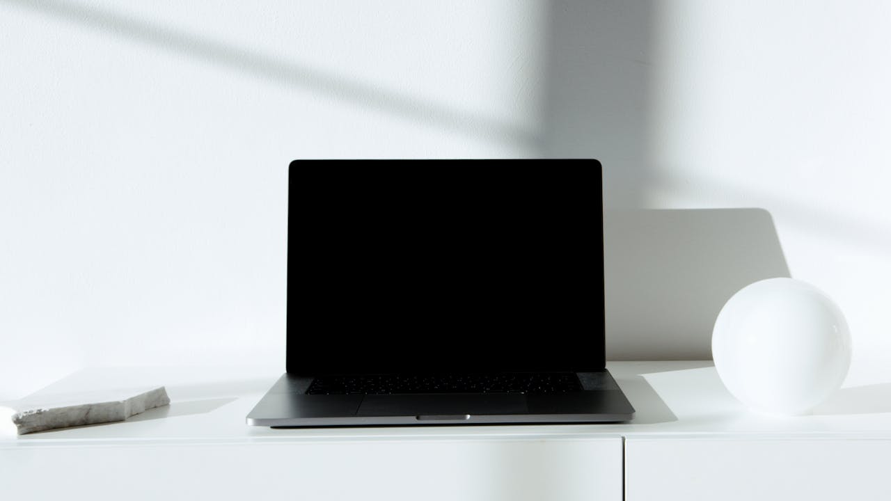 A sleek black laptop on a white desk in a minimalist setup with natural lighting.