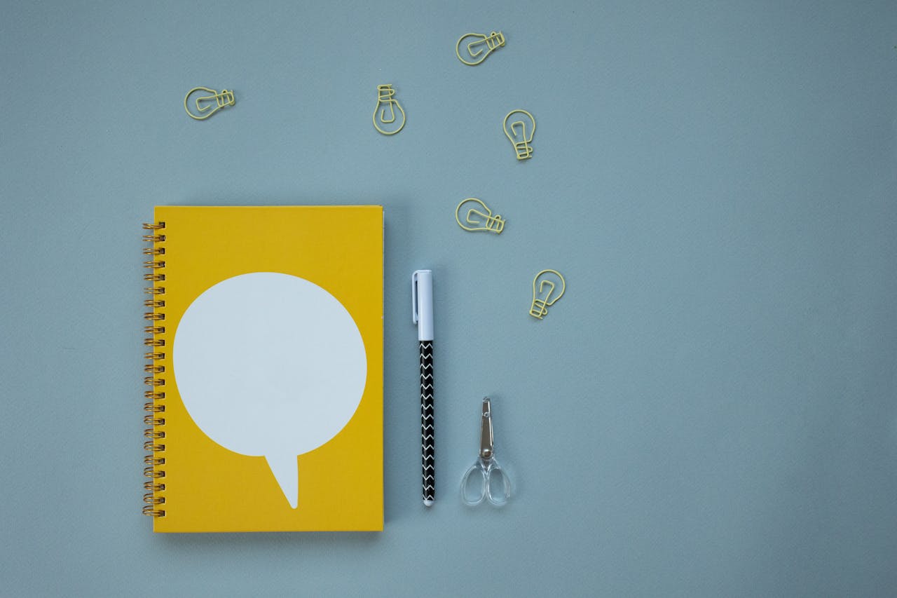 Creative workspace setup with a yellow spiral notebook, pen, scissors, and light bulb paper clips on a blue background.