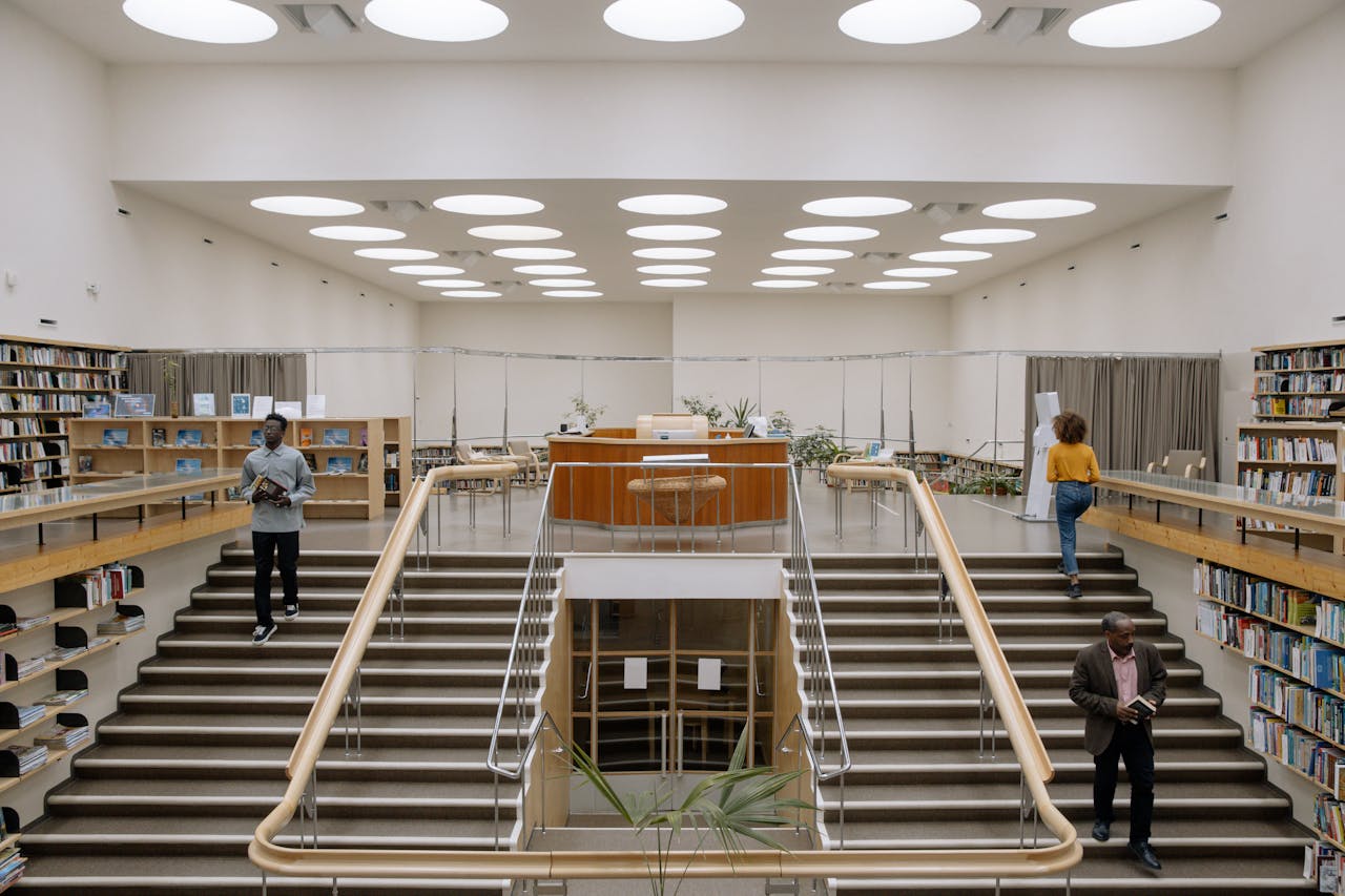 Spacious library interior with modern design, bookshelves, and people on stairs.