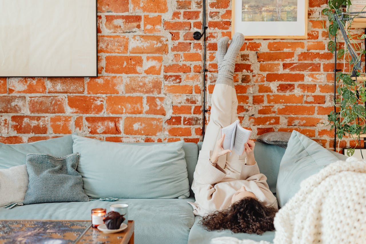 Woman relaxing on a sofa with book against brick wall, creating a cozy indoor vibe.